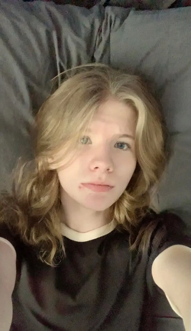 Sissy twink with blonde hair and pale skin lying on a grey pillow in a black t-shirt