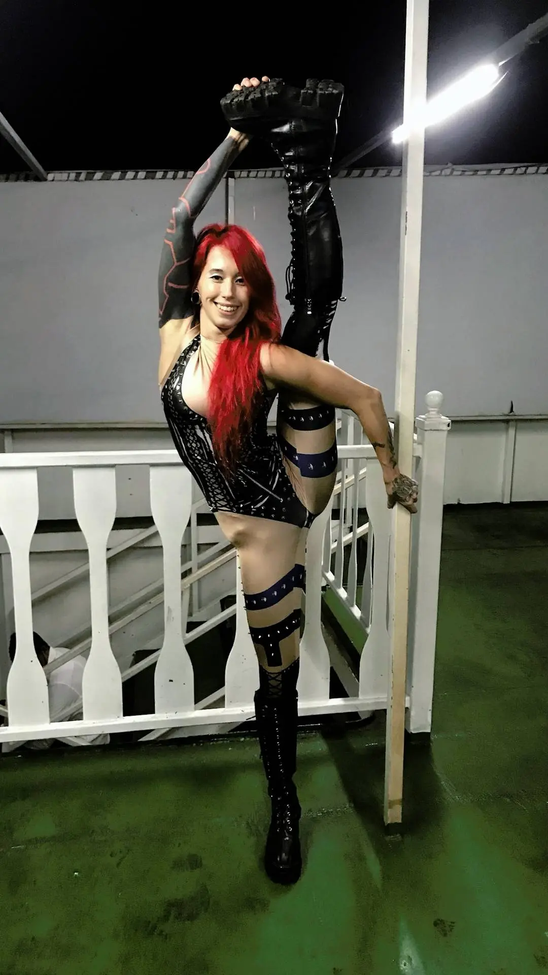Athletic Asian redhead in a black cutout bodysuit and thigh-high boots performing a high leg stretch outdoors
