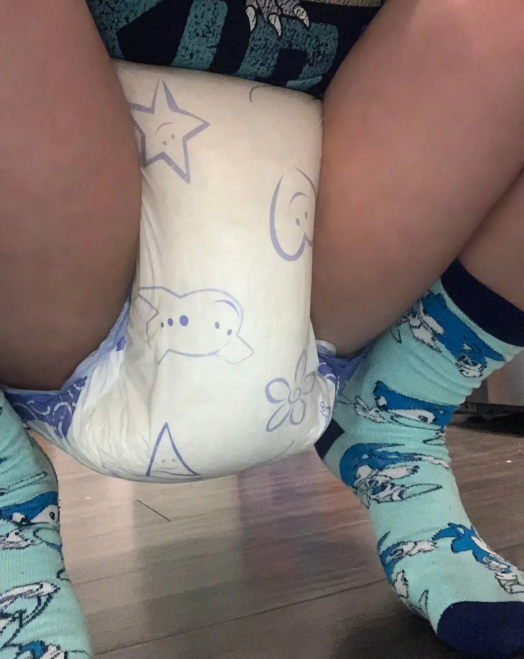 A curvy white person in a patterned adult diaper and Sonic the Hedgehog socks standing on a wooden floor.