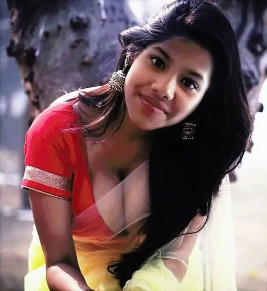 Busty Indian bhabhi in a red blouse and yellow saree leaning forward and smiling outdoors