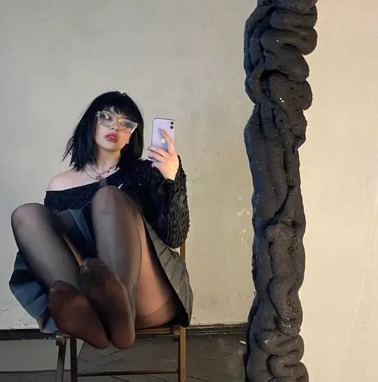 Goth girl in a black off-the-shoulder top, grey mini skirt, and black sheer nylon tights taking a mirror selfie