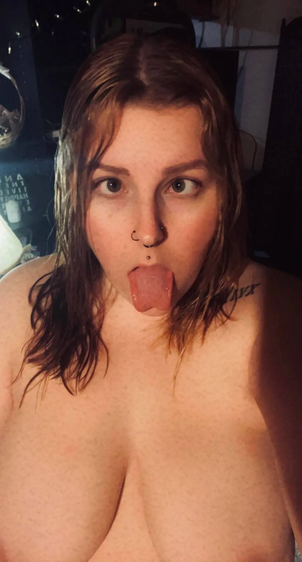 Busty brunette woman with a nose ring and tongue piercing sticking her tongue out in a naked selfie.