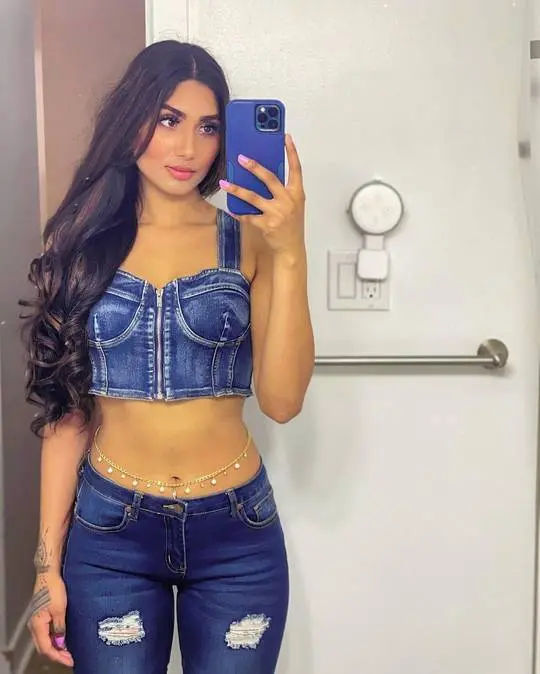 Curvy Desi American woman in a denim crop top and ripped blue jeans taking a mirror selfie with a gold waist chain.