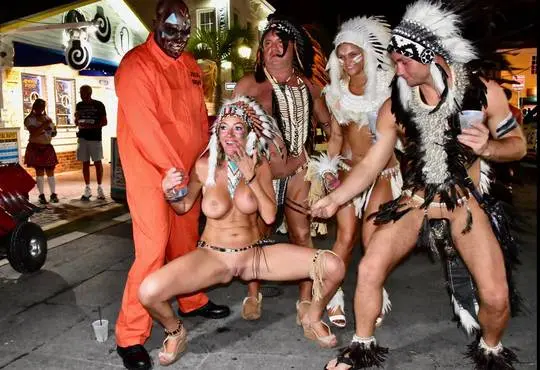 Interracial group in Native American costumes and an orange jumpsuit posing nude at a wild Halloween party.