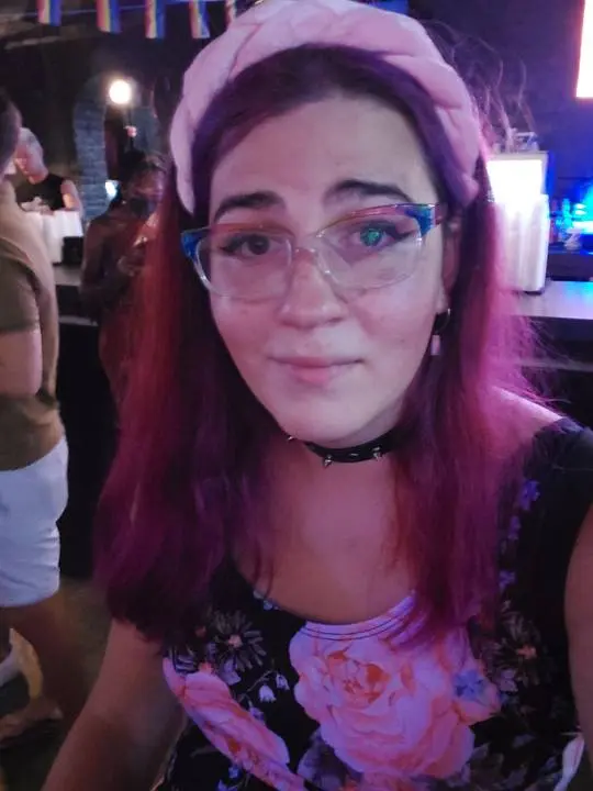 Trans girl with pink hair, glasses, a pink headband, and a floral top posing for a selfie at a gay bar.