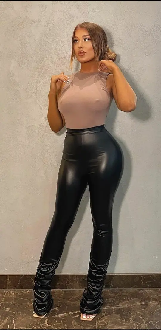 Eve Gale posing in a beige sheer top and black shiny faux leather flare pants against a grey wall