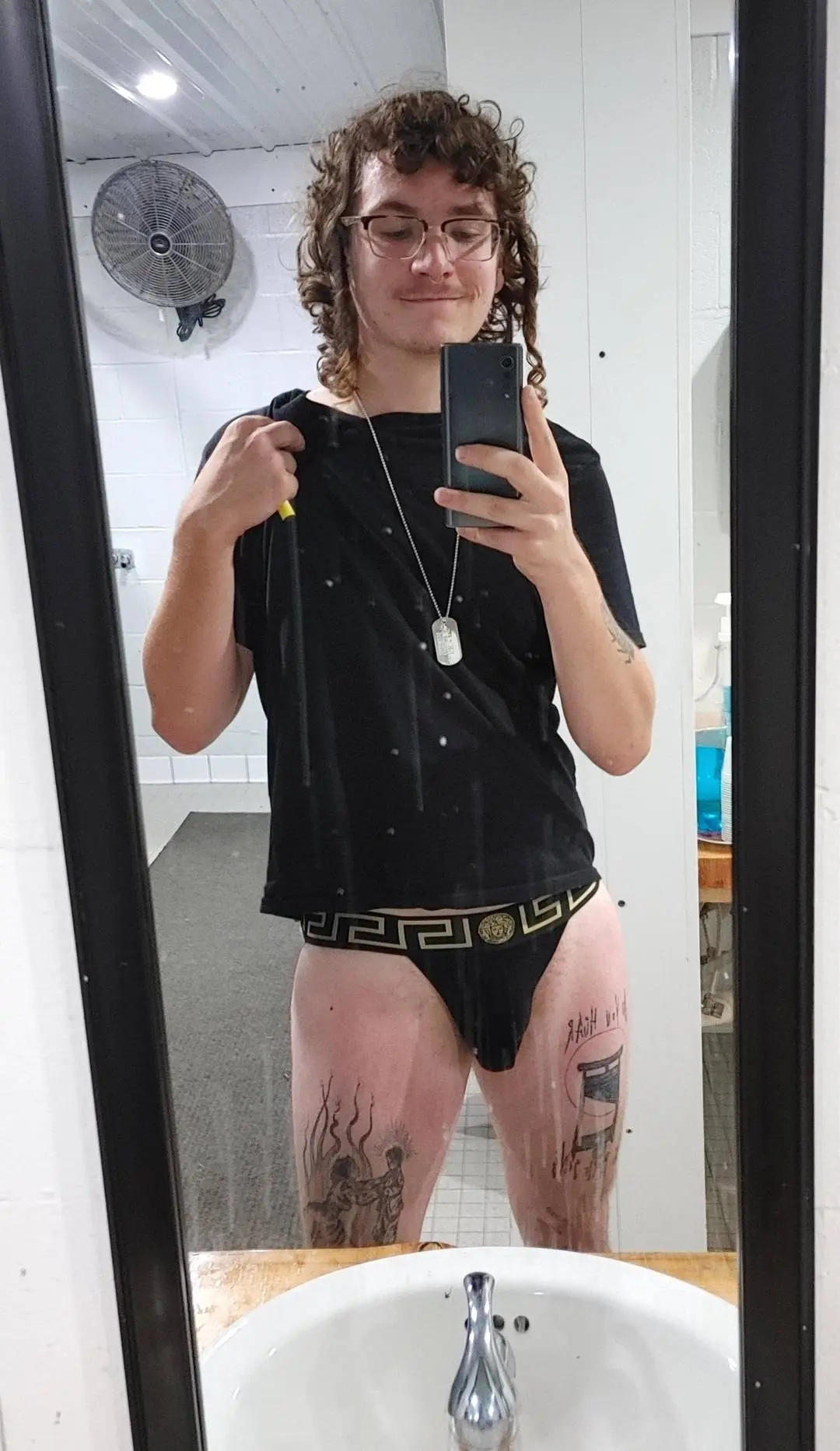 Slim white man with curly hair and tattoos taking a mirror selfie in a bathroom wearing a black t-shirt and underwear