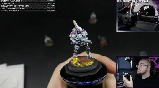 A person holding a painted Warcaster miniature figure next to an Ender 3 3D printer in a painting stream screenshot.