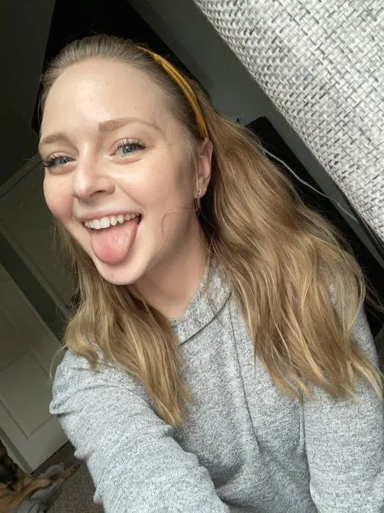 Blonde woman in a grey sweater and yellow headband sticking her tongue out in an ahegao selfie