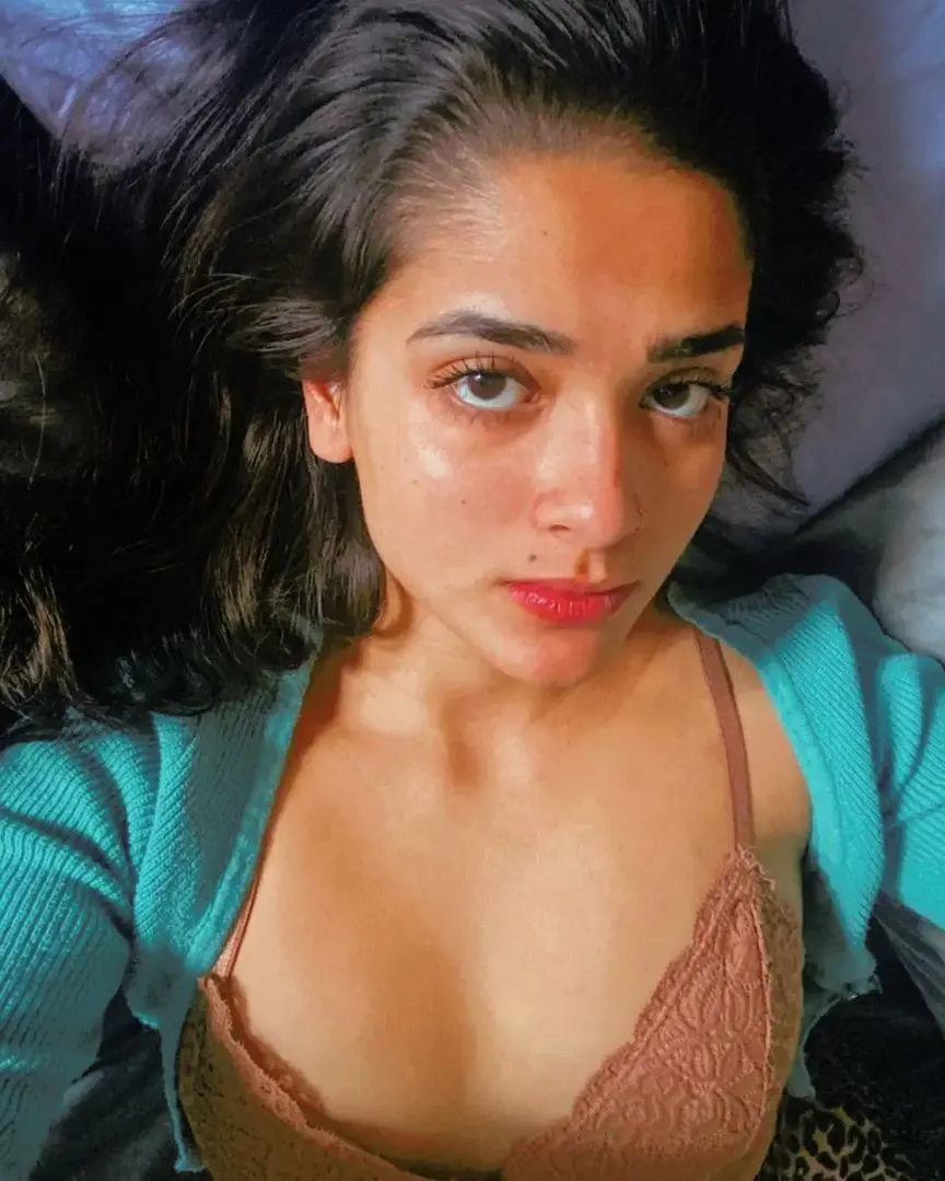 Anika Zulfikar, a busty desi girl with long dark wavy hair wearing a lace bra and teal cardigan in a bedroom selfie.