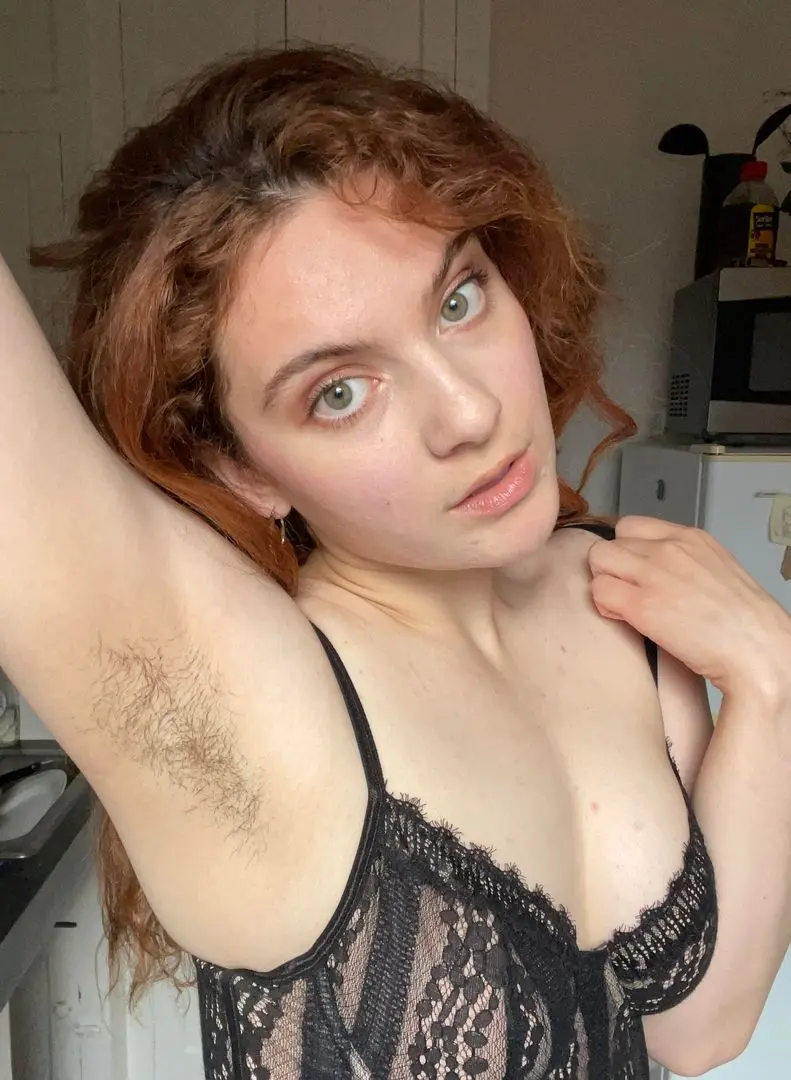 Slim white woman with curly red hair in a black lace camisole posing with arms up to show her hairy armpits
