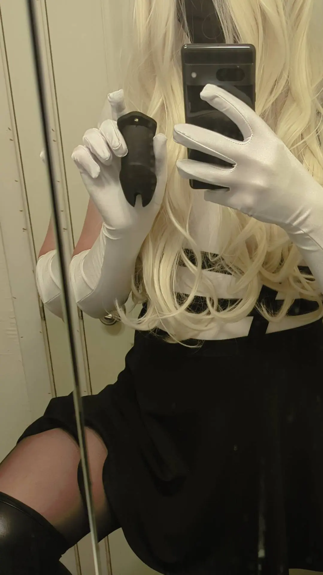 Femdom woman in a blonde wig, white gloves, and black skirt taking a mirror selfie with a dildo