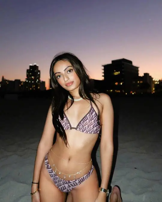 NRI American Gujju beauty in a patterned bikini and gold body chain on a beach with a city skyline at sunset