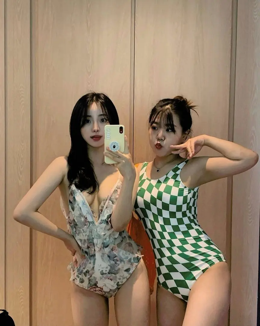 Two Korean women in one piece swimsuits, one floral and one green checkered, posing for a mirror selfie.