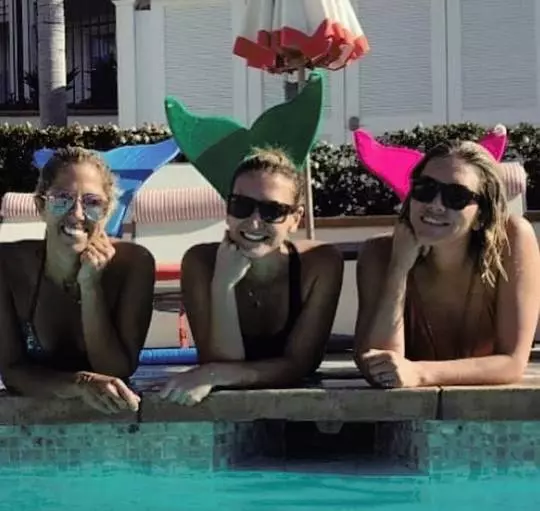 Sara Walsh, Cynthia Freeland, and Charissa Thompson in swimwear by a swimming pool with mermaid decorations