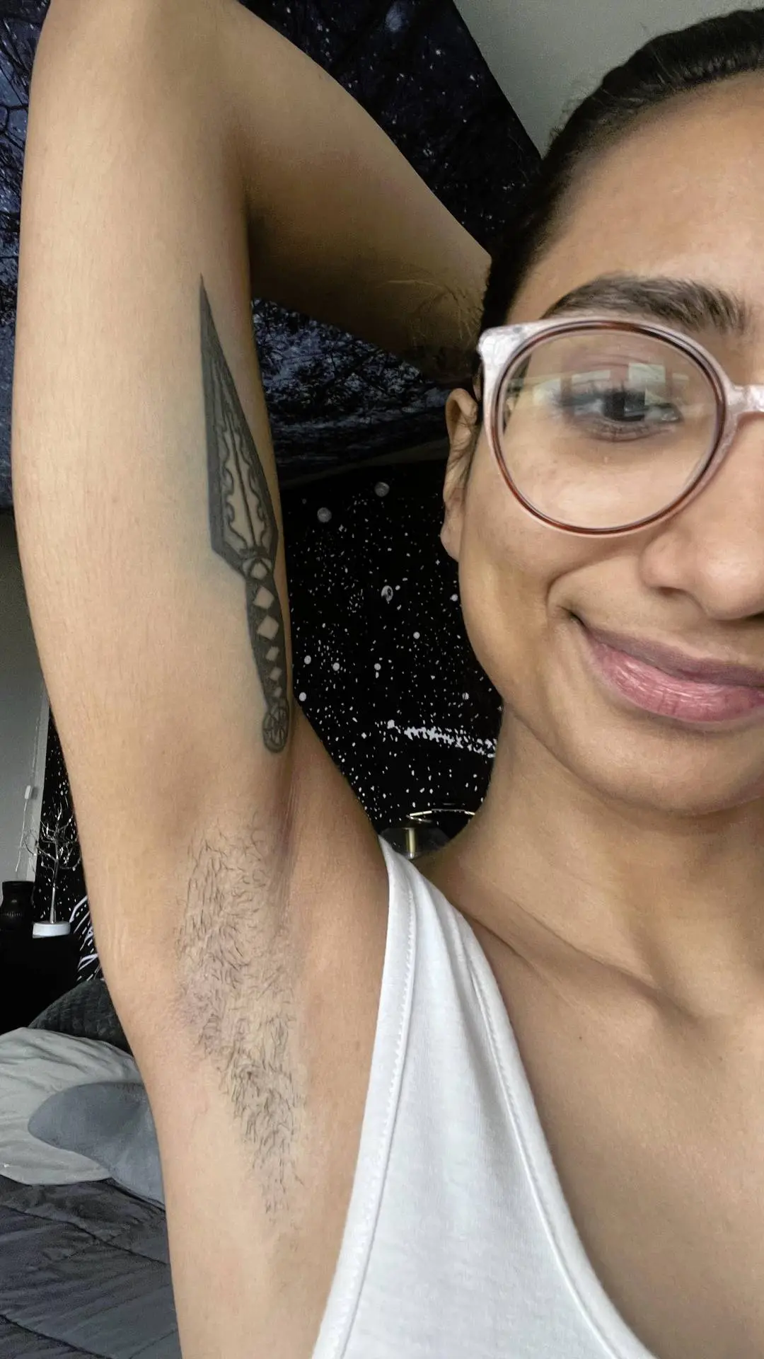 shagisabadword, a slim Indian woman with glasses and a white tank top, posing with hairy armpits and a dagger tattoo
