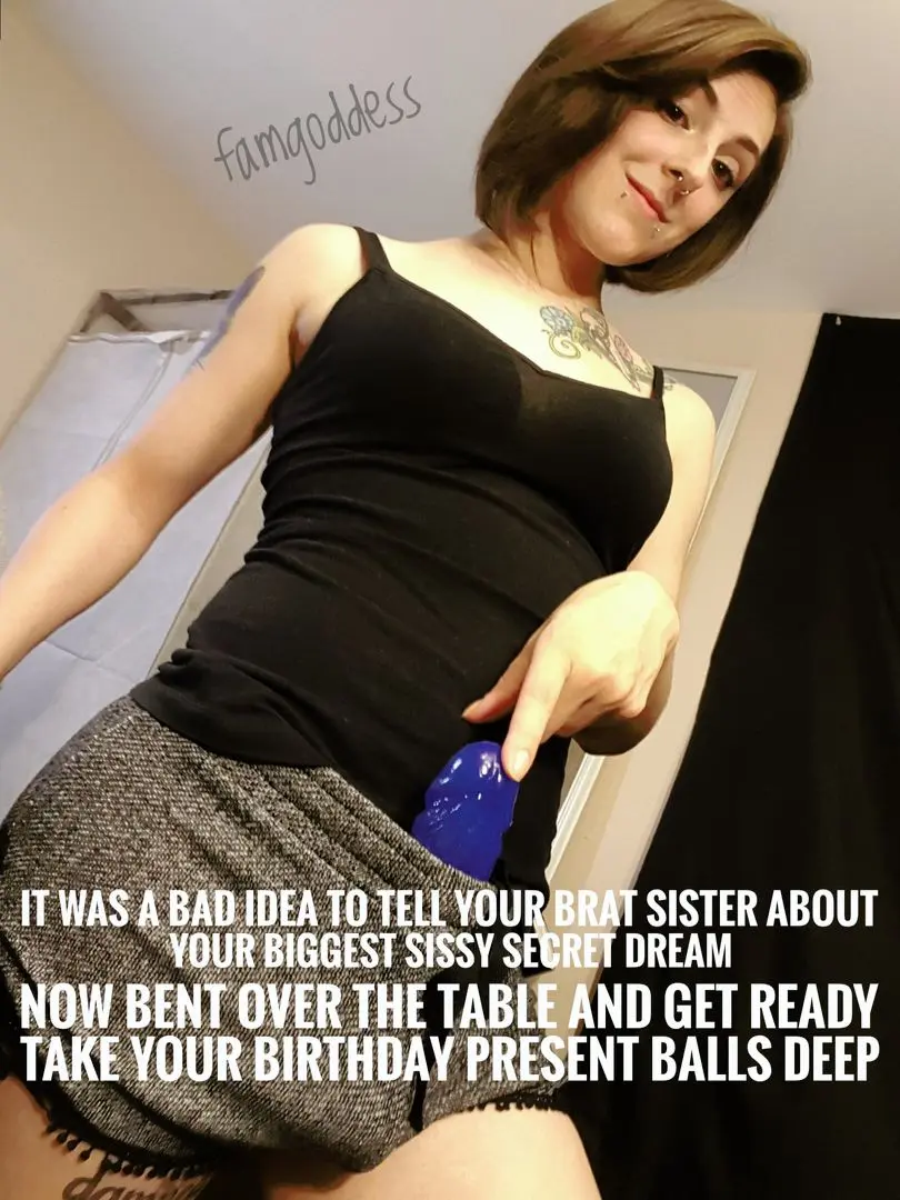 famgoddess in a black tank top and grey skirt holding a blue dildo with sissy and pegging captions