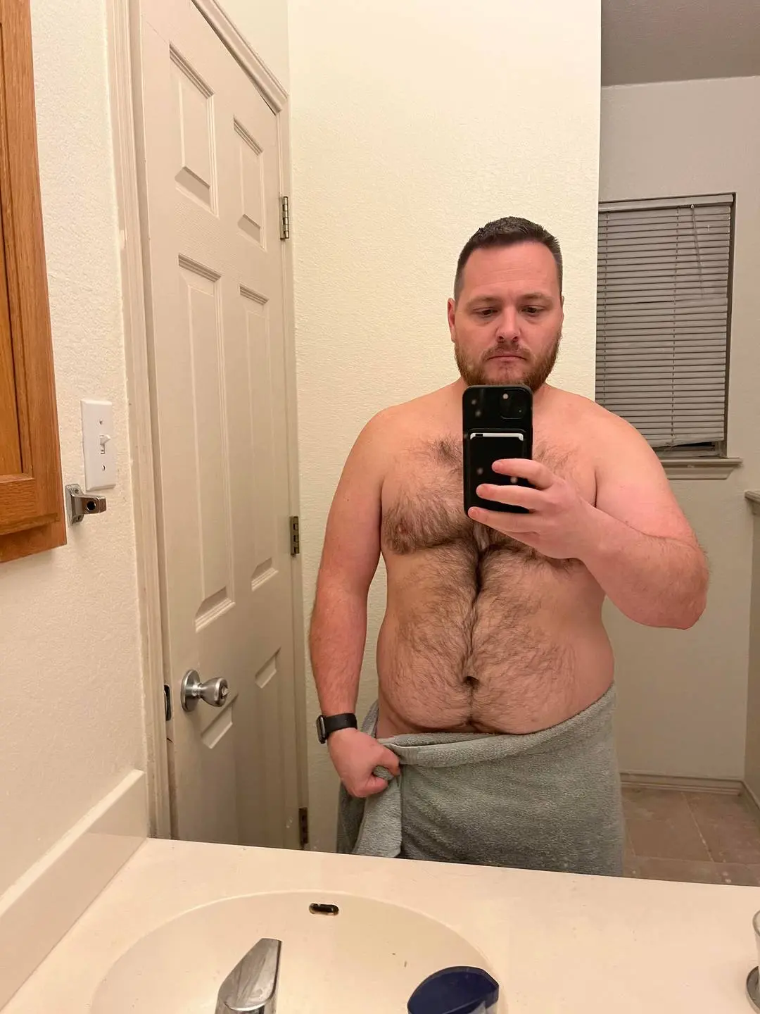 Shirtless white man with a hairy chest and belly wearing a grey towel in a bathroom mirror selfie
