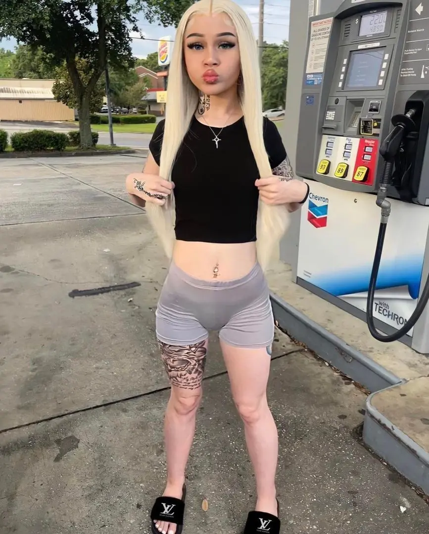 Ghetto white girl with long blonde hair and tattoos wearing a black crop top and grey shorts at a gas station