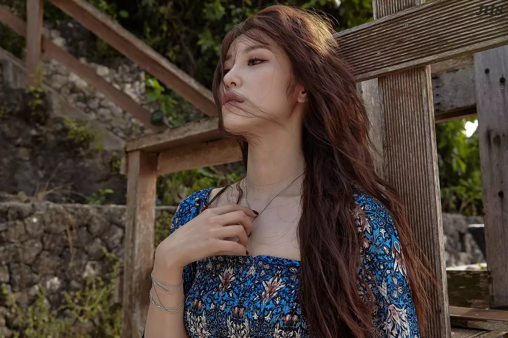 Jun Hyo Seong wearing a blue floral print sundress posing by a wooden structure and stone wall in Bali