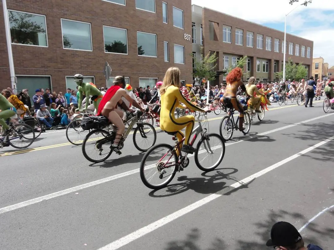 Slim brunette woman with yellow body paint riding a bicycle at a street fair, topless and nude