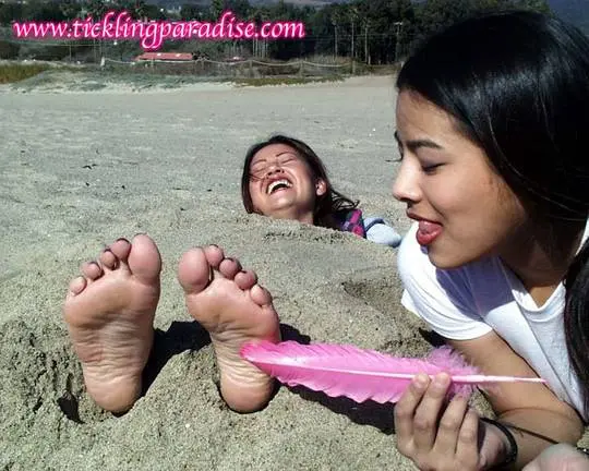 Asian women tickling feet and toes with a pink feather while buried in the sand at the beach