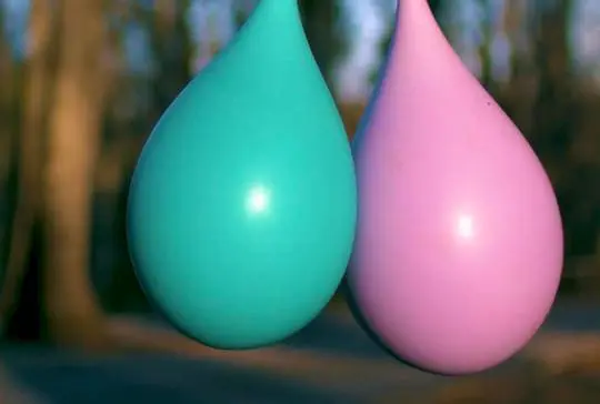 Two teardrop-shaped water balloons, one turquoise and one pink, hanging side by side in a blurred forest.