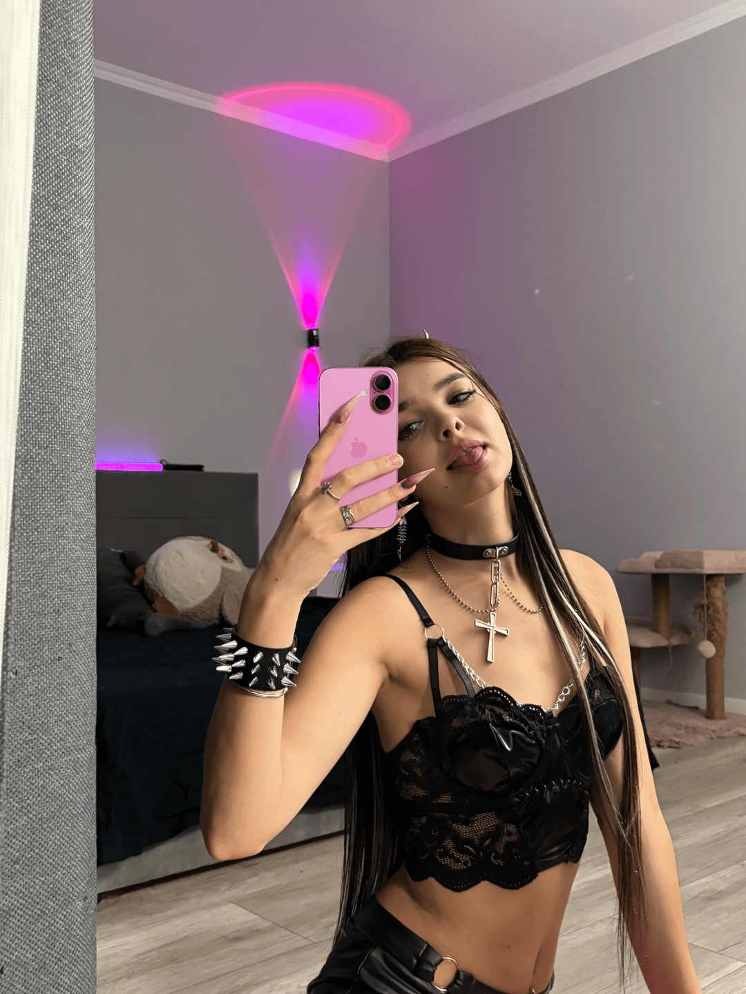 E-girl with long brown hair in a black lace crop top and leather pants taking a mirror selfie with her tongue out.