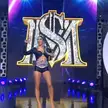 Mercedes Moné with blue hair in a black leather cropped top and shorts wearing championship belts