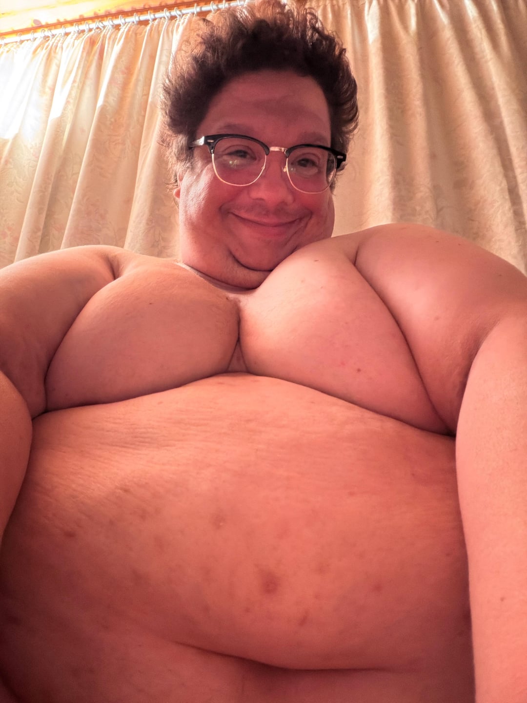 Shirtless white bbw man with curly hair and glasses smiling for a selfie in front of curtains.