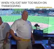 A heavy man in a white polo shirt rubs his stomach and throws his arms up in a broadcasting booth at a baseball stadium.