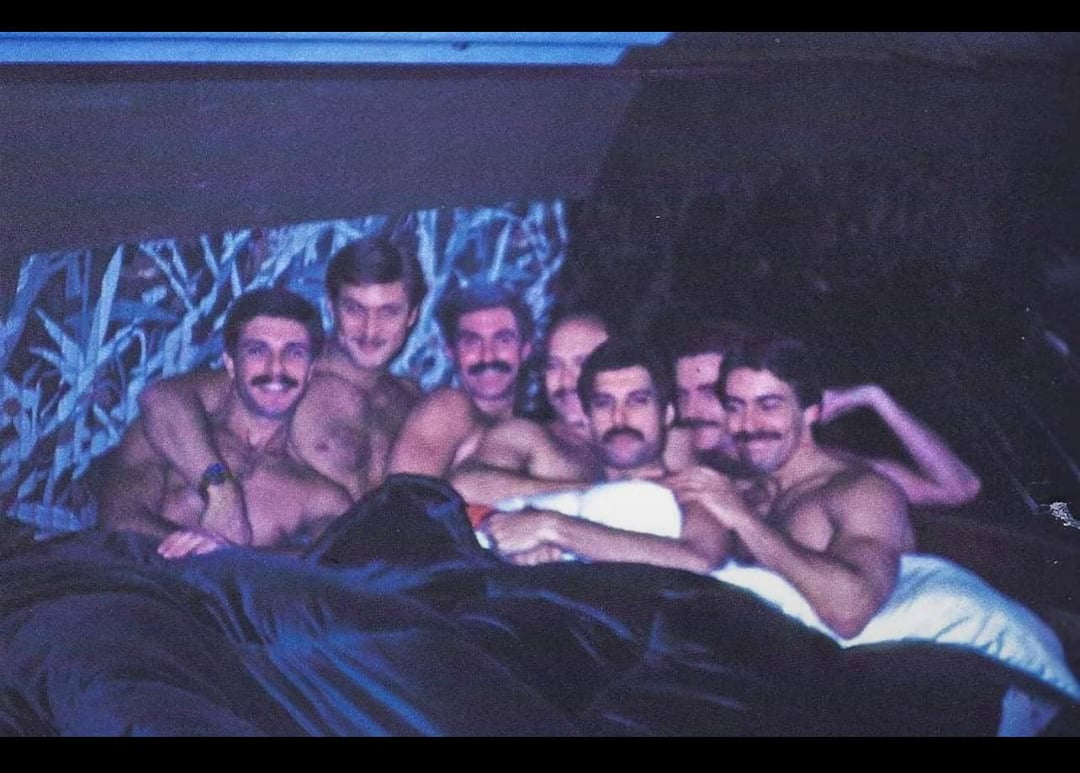 Freddie Mercury and a group of shirtless men with mustaches lying under blankets in a bed