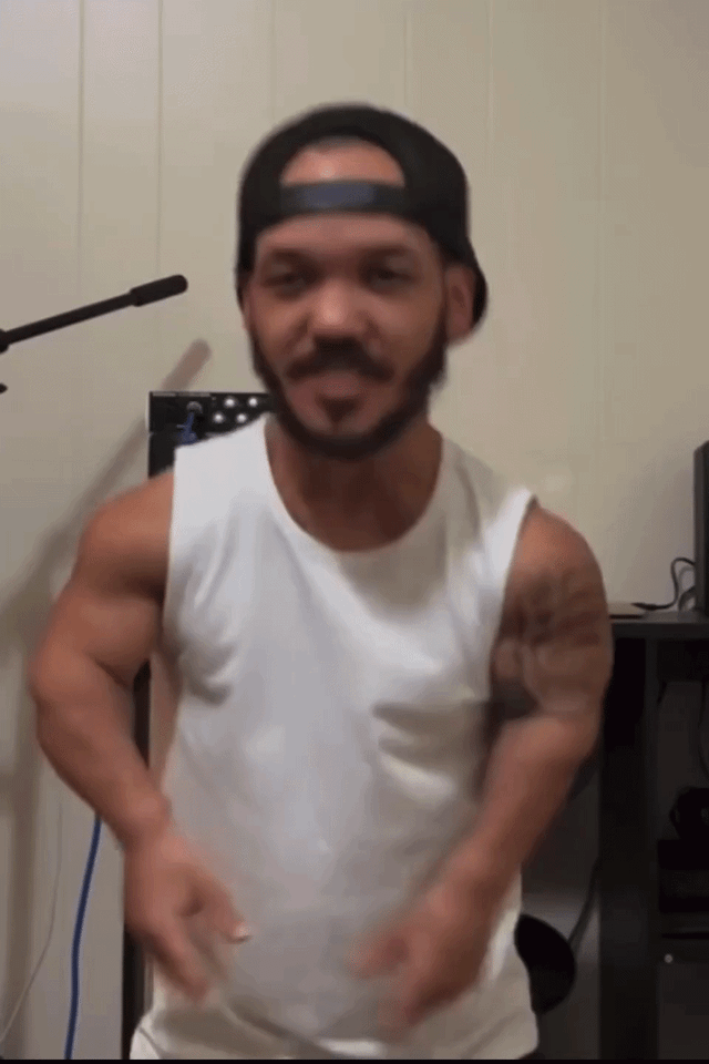 A muscular man with a tattoo and black baseball cap dancing in a white tank top