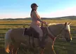 Curvy white brunette cowgirl riding a horse naked in a field wearing a riding helmet and boots