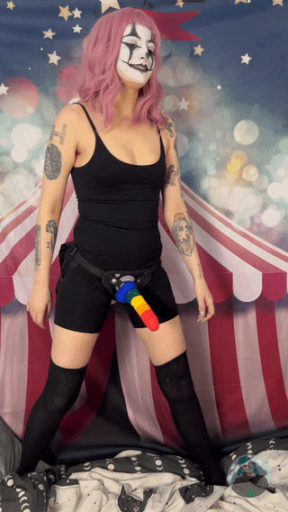 Hairy strap-on girl with pink hair in clown makeup and a huge dildo posing against a circus backdrop