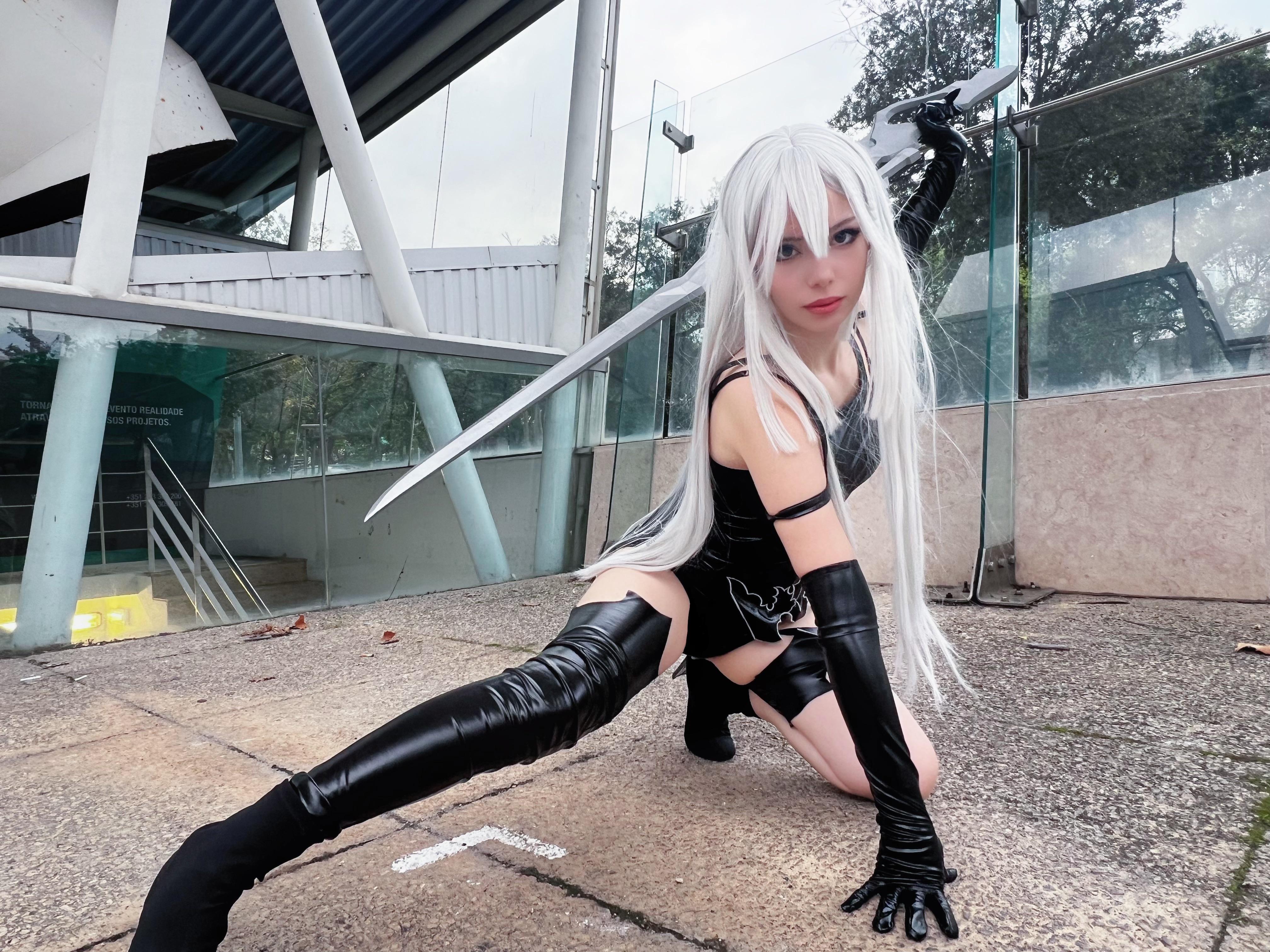 A2 cosplay in a black bodysuit and thigh-high boots posing with a sword by a glass wall