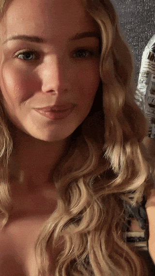 Julia Beautx smiling and winking in a selfie POV clip, featuring her long blonde curly hair and black top.