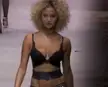 Rose Bertram walking a fashion show runway in black lingerie, showing her curly blonde hair and tanned skin.