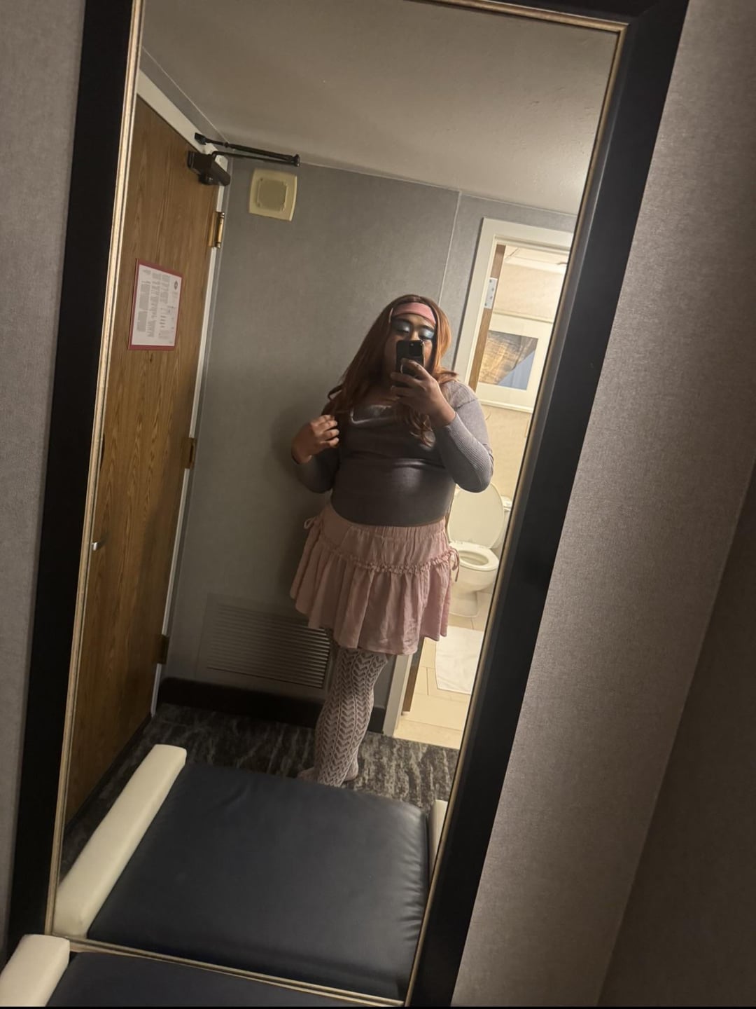 Curvy redhead sissy in a grey long-sleeved top and pink ruffled mini skirt taking a mirror selfie in a bathroom