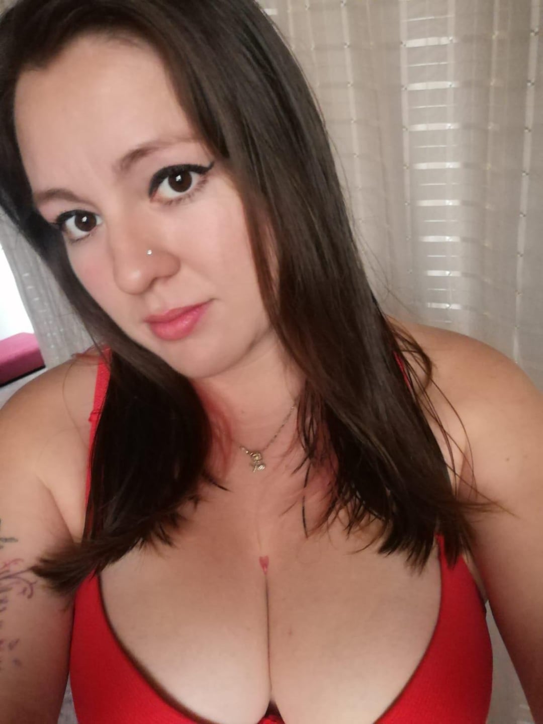 Busty white woman with long brown hair, a nose piercing, and arm tattoo wearing a red top in a selfie.