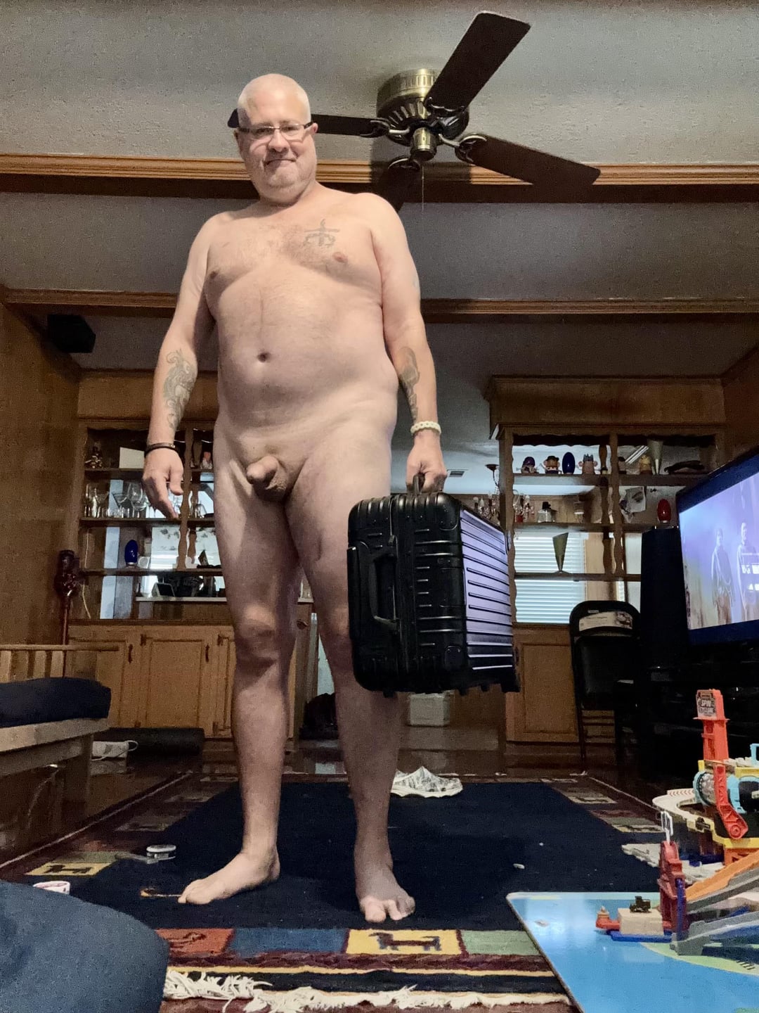 Mature bald white man with tattoos standing naked in a living room holding a black suitcase.