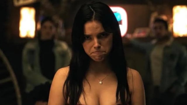 A woman with long dark hair and a red v-neck top making a funny face with blurred men in the background.
