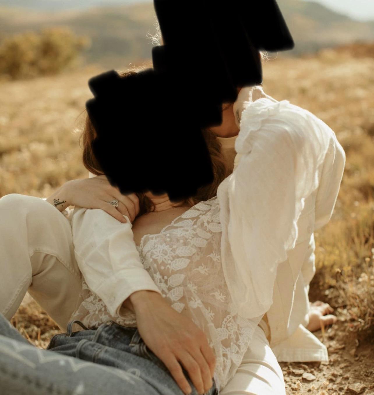 Woman in a white lace blouse and blue jeans embracing a man in an autumn field with mountains.