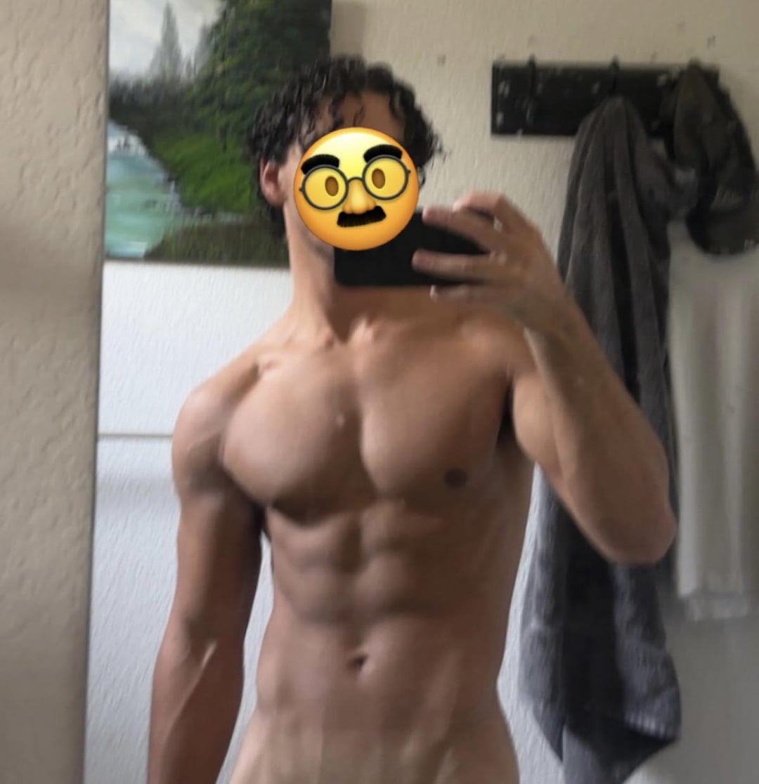 Muscular shirtless man with curly hair taking a mirror selfie in a bathroom, fit and tanned.