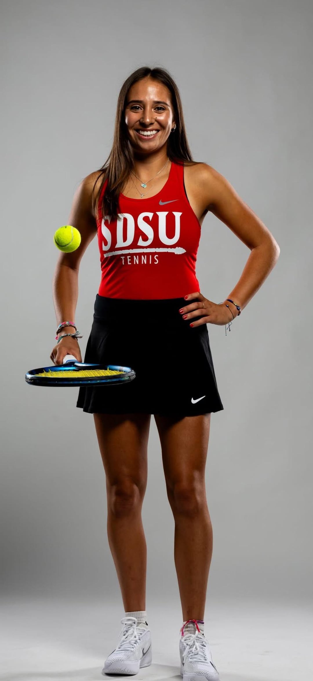 Smiling athletic latina in a red SDSU tennis tank top and black short skirt holding a racket and ball