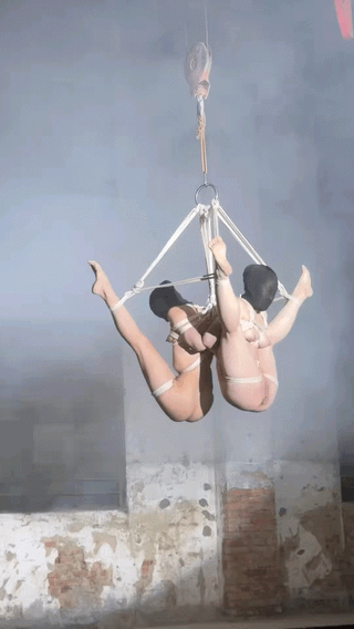 Two slim Asian women in white lingerie suspended in shibari rope bondage in an industrial room.
