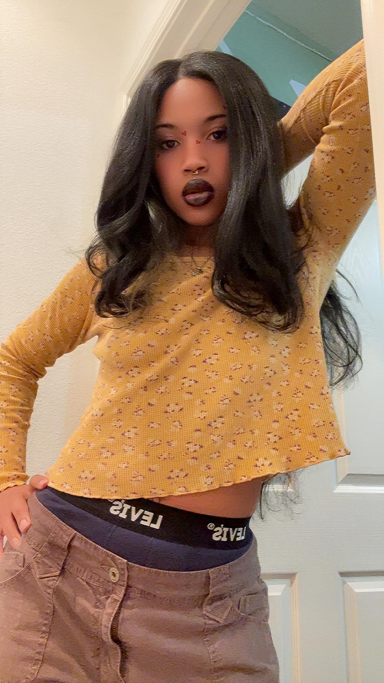 Mixed black girl with long black wavy hair in a yellow crop top and brown pants showing Calvin Klein underwear
