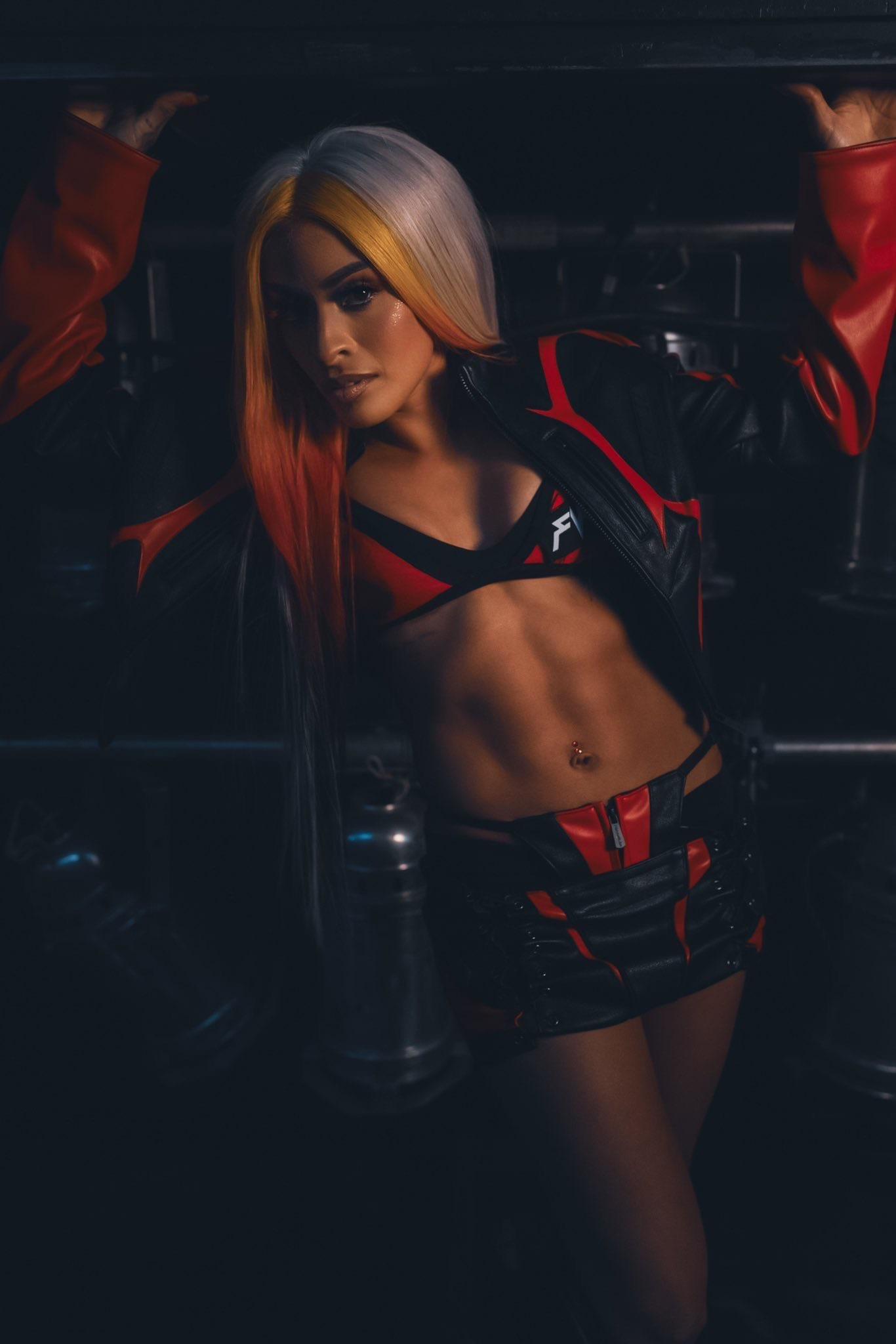 Zelina Vega in a red and black leather jacket, sports bra, and mini skirt posing in a dark industrial setting