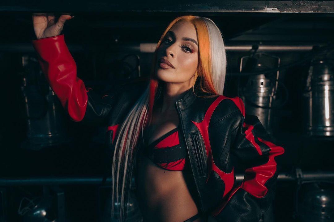Zelina Vega in a red and black leather jacket, sports bra, and mini skirt posing in a dark industrial setting