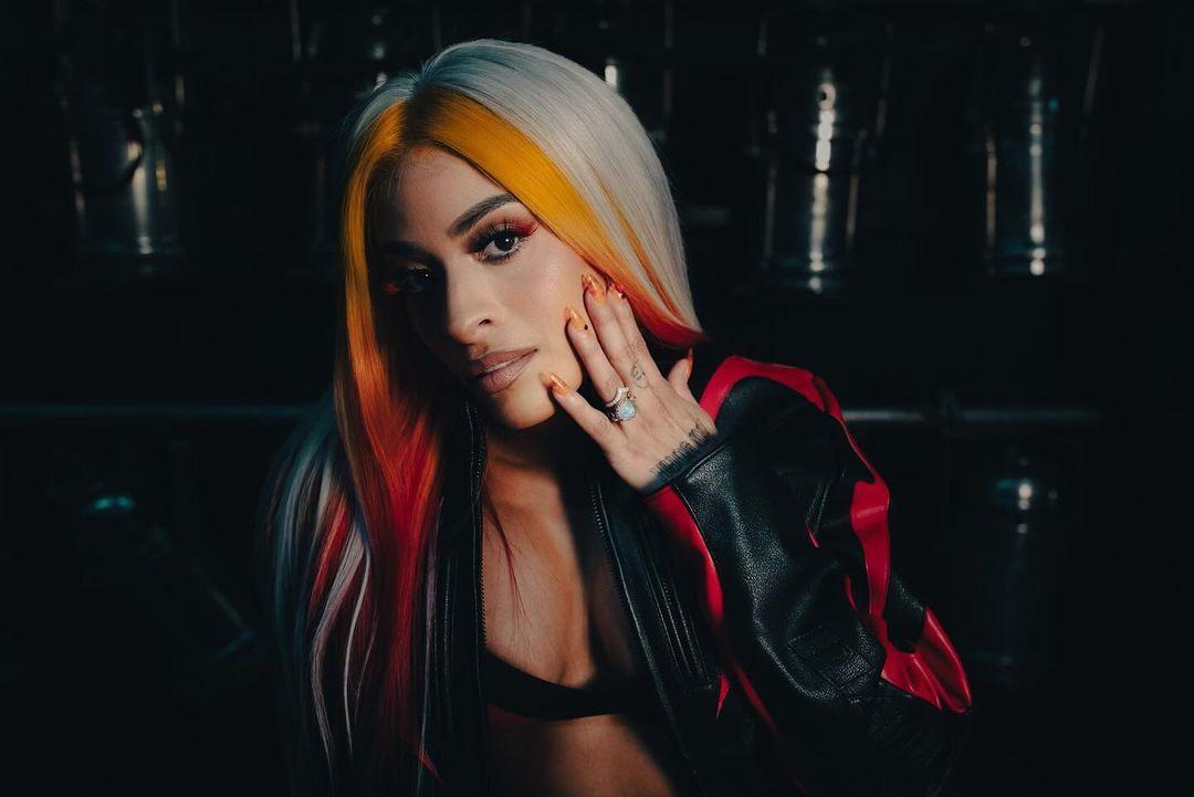 Zelina Vega in a red and black leather jacket, sports bra, and mini skirt posing in a dark industrial setting